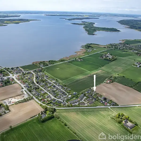 Landsby ved sø grænser op til grønne marker, der strækker sig ud mod horisonten. Boliger ligger samlet i bebyggelse nær hovedvej, med en stor sø og skove i baggrunden.