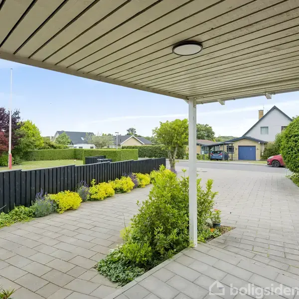 Overdækket terrasse med flisebelægning strækker sig ud til en have med grønne buske og blomster, omgivet af sorte træhegn i et villakvarter med huse i baggrunden.