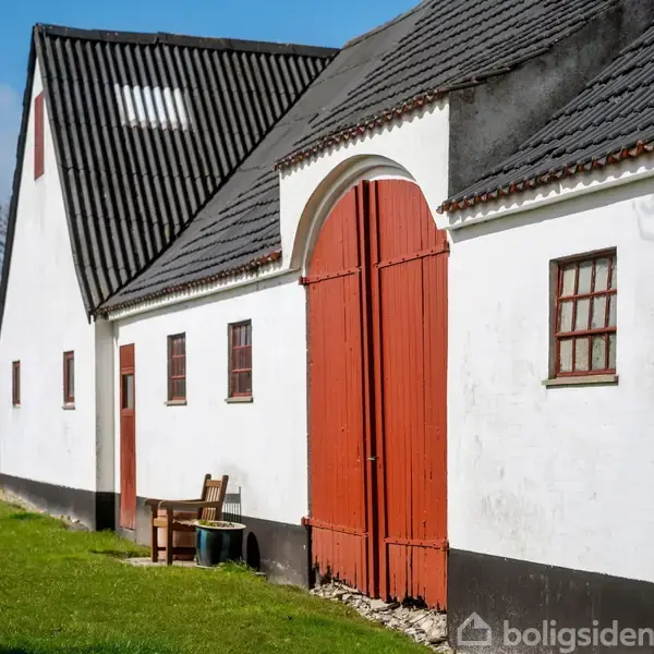 En hvid bygning med sorte tage og røde trædøre står i en grøn have. En bænk er placeret ved siden af dørene. Skyer ses på den blå himmel.