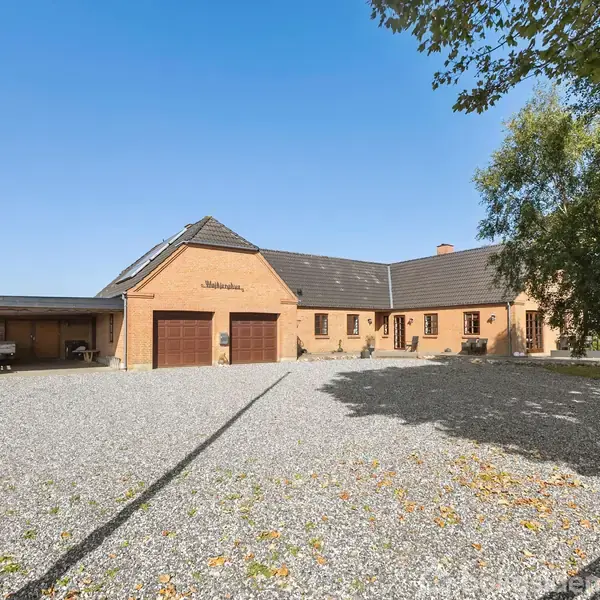 En orange murstensbygning med to garageporte, teksten "Stærdalen" på facaden. Bygningen står på en gårdsplads belagt med grus, omgivet af træer og marker under en blå himmel.