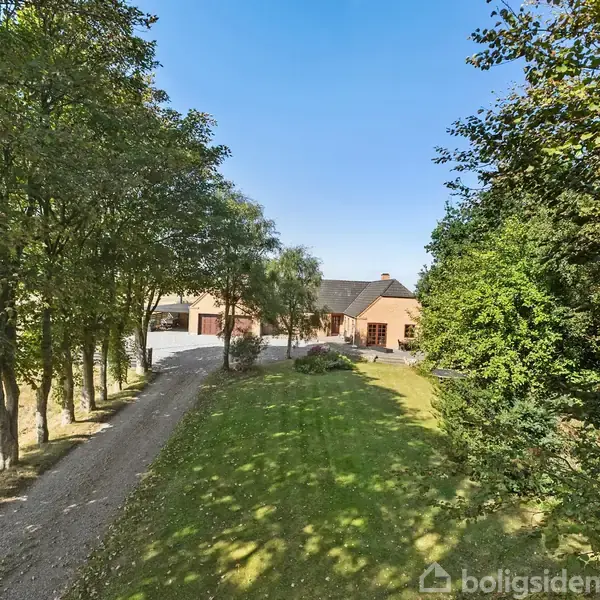 En grusvej fører mod et hus omgivet af træer og grønt græs. Huset har en jordbrugsstil og er beliggende i en landlig have.