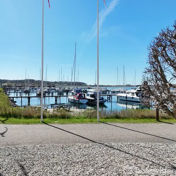 Både ligger stille ved en marina. Folk går langs en sti, og omgivelserne består af græs og træer. Himmel er klar og blå.