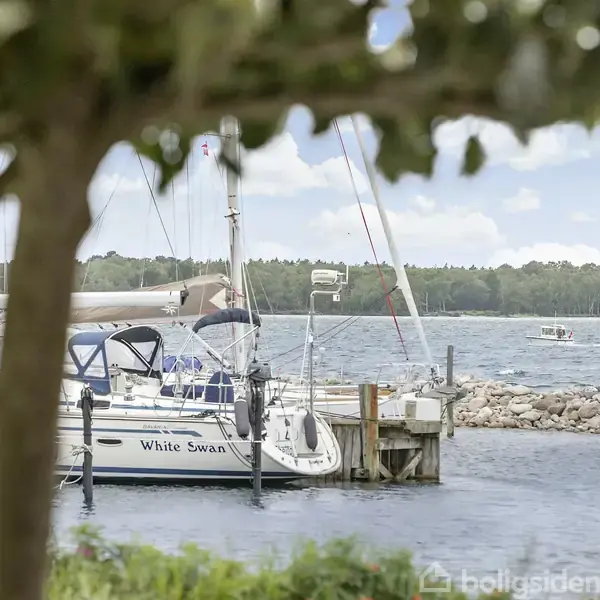 Sejlbåd med navnet "White Swan" ligger til kaj ved en havn, omgivet af roligt vand og trærige omgivelser. Skyer fylder en del af den klare, blå himmel.