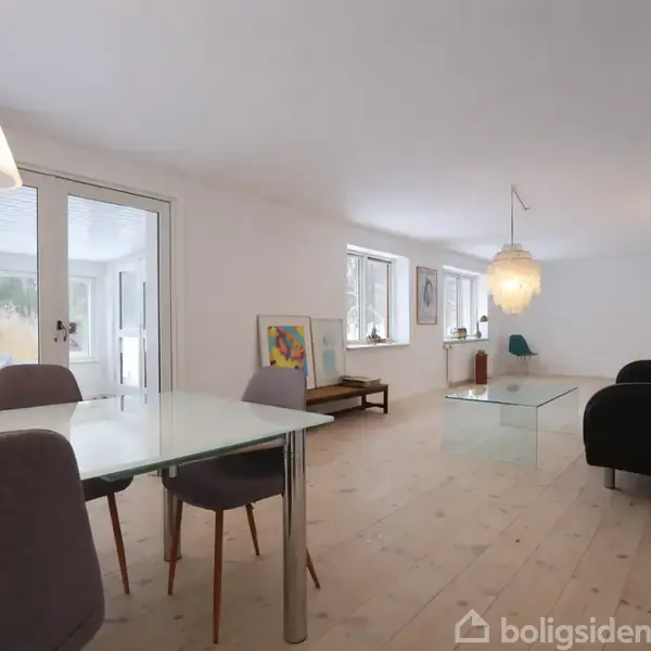 Et moderne spiseområde med et glasbord og stole står i en lys stue. Store vinduer lader naturligt lys strømme ind, mens en sort sofa og lamper skaber en hyggelig atmosfære.