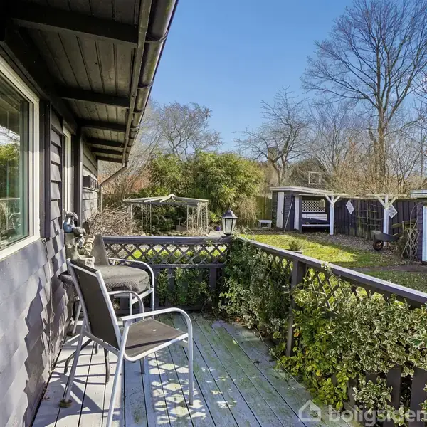 Terrasse med sorte stole og borde står ved siden af et sortmalet hus. I baggrunden ses en have med græsplæne, træer og en mindre bygning i træ.