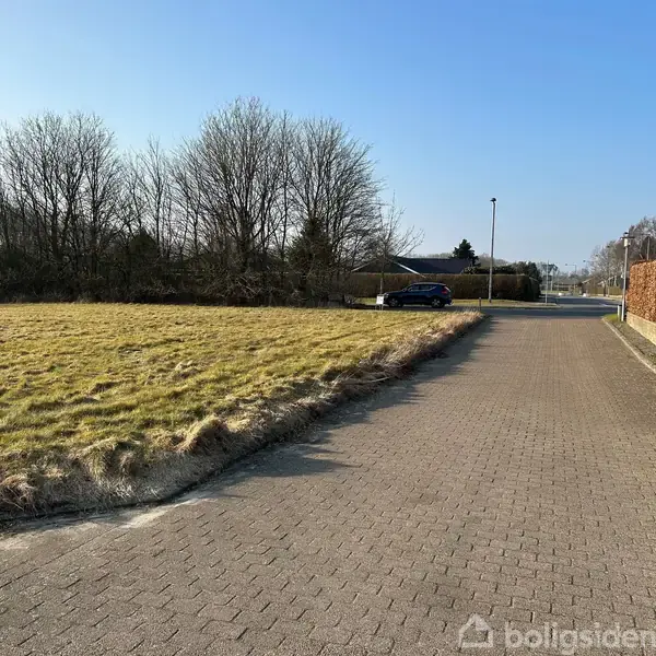 En bil holder stille på en brolagt vej. Til venstre er en græsmark, og til højre en hæk. Baggrunden domineres af træer under en skyfri himmel.