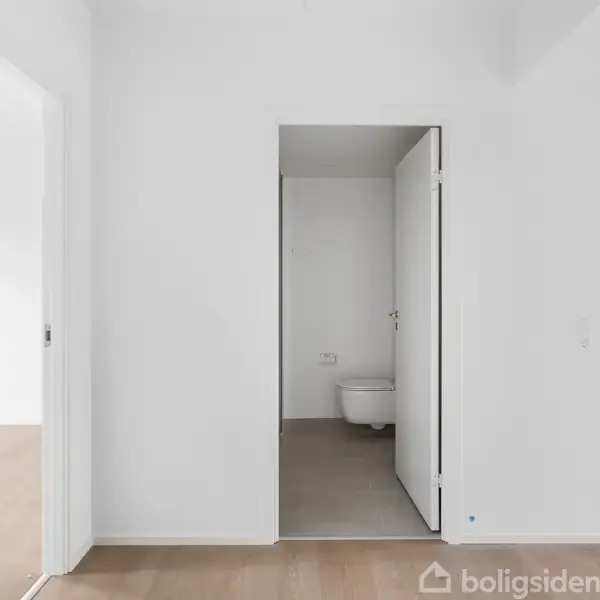 Dør åbner til et badeværelse med toilet, omgivet af hvide vægge og lyst trægulv. Venstre side åbner til et andet rum med samme gulv.