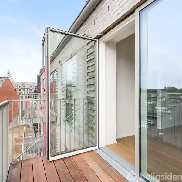 En åben altandør fører ud til en træbelagt balkon, med udsigt over en moderne bydel med byggedelejlighedskomplekser og tage.