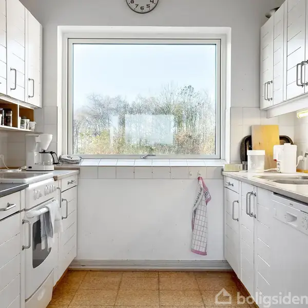 Et lyst køkken med hvide skabe er indrettet med ovn, kaffemaskine og vask. Et stort vindue slipper naturligt lys ind, og der hænger et viskestykke ved vasken.