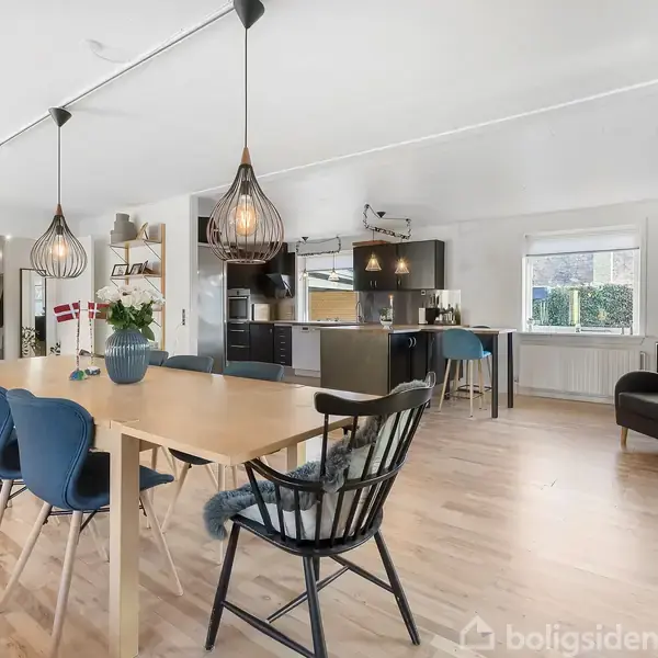 En stor spisestue med et lyst træbord og blå stole, placeret i et åbent køkkenområde. To hængelamper lyser rummet op. Grønne planter dekorerer området.