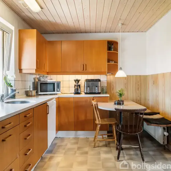 Køkken med træskabe, bordplade og vask ved vindue. Mikroovn og brødrister står på hylden. Spisebord med stole og bænk i hjørne. Lyse fliser og træpaneler.