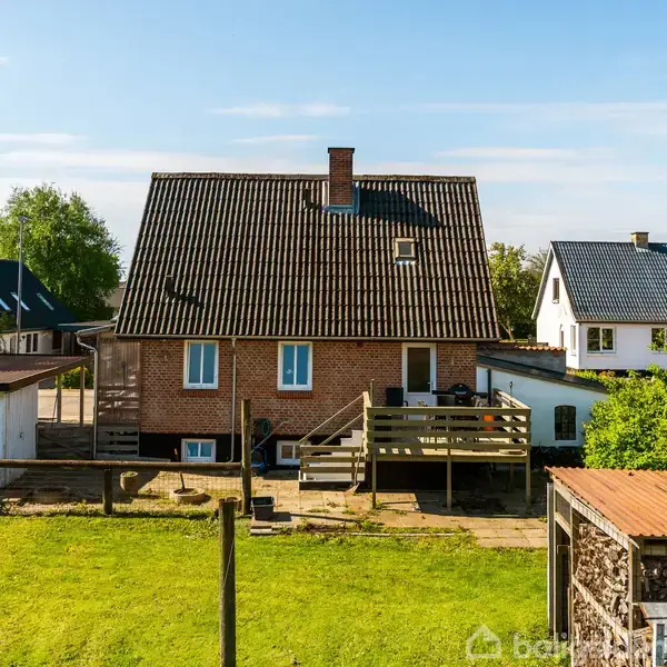 Rødt murstenshus med sort tag står stille. Træterrasse foran huset, omgivet af grønne haver og flere mindre bygninger. Blå himmel og andre huse i baggrunden skaber en stille nabolagsstemning.