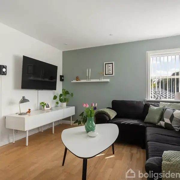 Sort hjørnesofa står i en lys stue, dekoreret med grønne puder og et sofabord. På væggen hænger et TV over en hvid skænk. Et vindue giver naturligt lys.