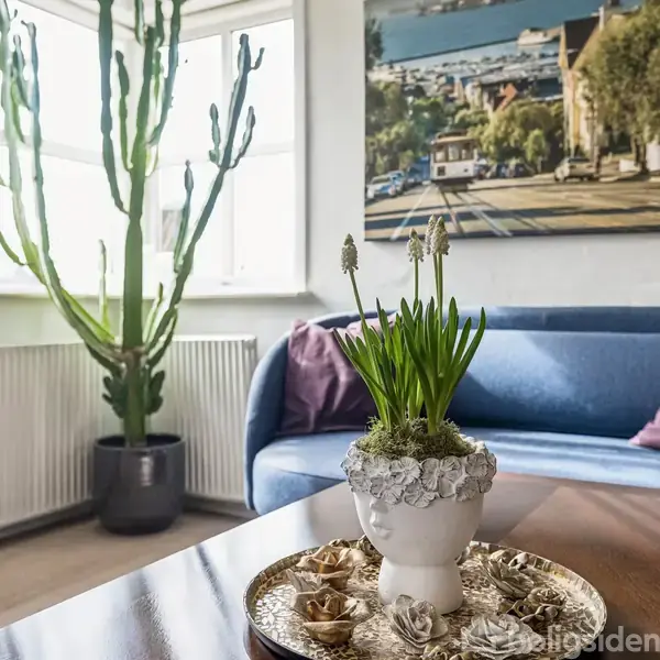 En potteplante står på et sofabord med dekorative elementer, mens en stor kaktus ses ved et vindue. En blå sofa og et maleri dominerer baggrunden i en lys stue.
