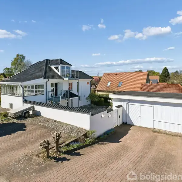 Et moderne, hvidt to-etagers hus med sort tegltag står i en forstadsområde. Foran er en flisebelagt indkørsel og en garage. Omkringliggende haver er velplejede med enkelte træer.