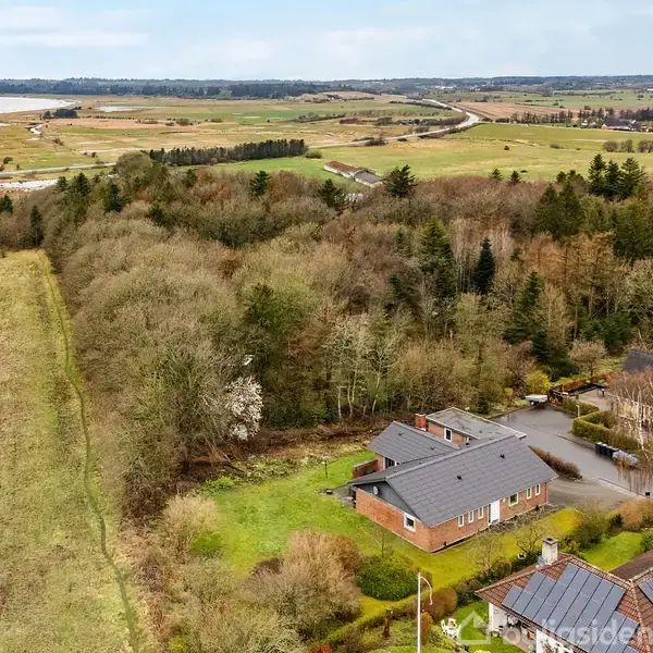 En enkeltstående murstensvilla ligger blandt træer ved en skovkant. Omkring huset er en stor have. I baggrunden ses åbne marker og en vej, der snor sig gennem landskabet.