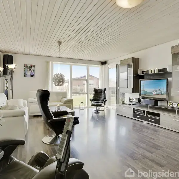 Stue med hvide sofaer og sort lænestol, centreret omkring et fjernsyn på et moderne TV-møbel. Store vinduer slipper naturligt lys ind, og gulvet er mørkt træ.