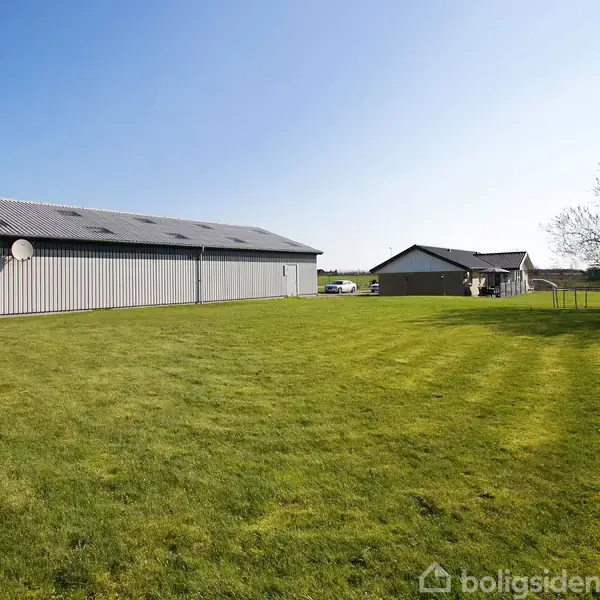 En stor lade står stille på en velholdt grøn græsplæne under en klar blå himmel. Til højre ses et mindre hus og nogle træer i baggrunden.