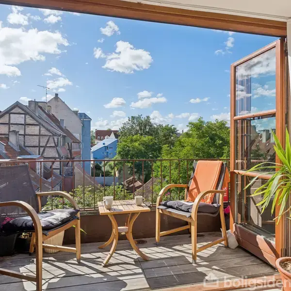 Balkon med to stole og et bord, der indeholder stearinlys, vender ud mod en byudsigt med træer og bygninger. Solskin fylder balkonen, omgivet af grønne planter.