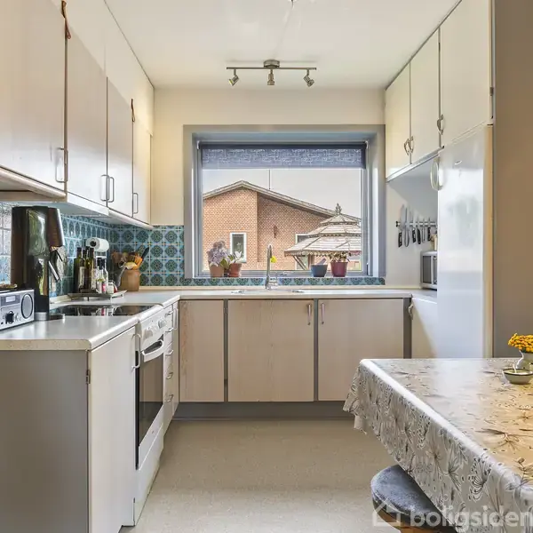 Køkken med hvide skabe og blå flisedetaljer, indeholder komfur og køleskab. Bordet er dækket med en voksdug og en blomstervase. Udsigt til hus gennem vindue.