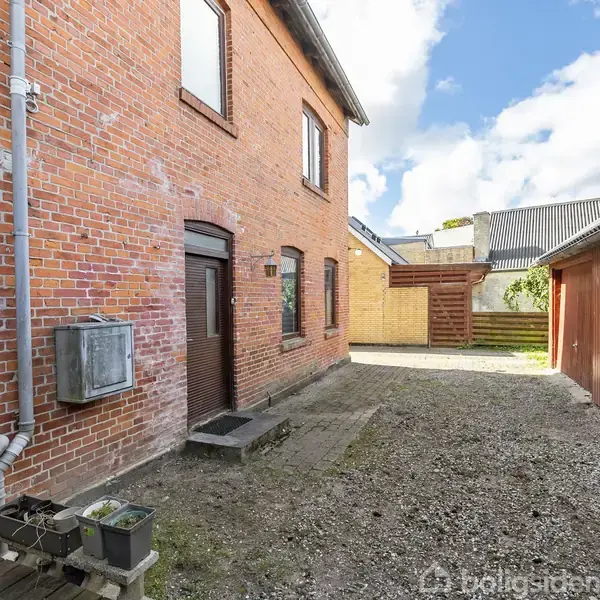 Rød murstensbygning med en brun dør står i en gårdsplads. På højre side er der en rød træbygning. Himlen er delvist skyet, og jorden er grusbelagt.