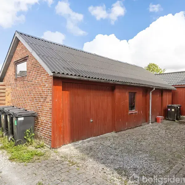 Bygning med røde trævægge og skråt gråt tag, står stille på flisebelagt område med skraldespande langs væggen. Blå himmel med hvide skyer i baggrunden.