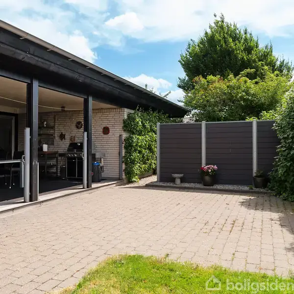 Terrasse med flisebelægning og åben overdækning til venstre, indeholdende spisebord og stole. En træhæk omkranser området, og en grå træhegn findes i baggrunden.