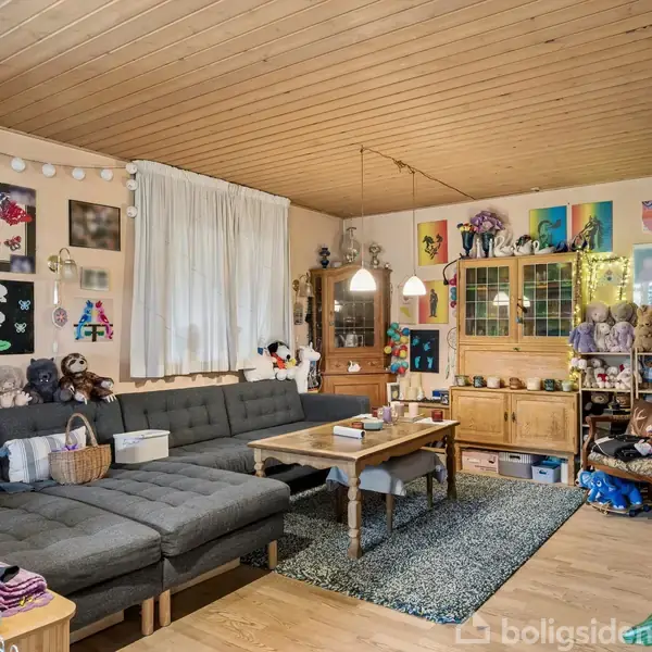 Grå hjørnesofa fyldt med pyntedyr og puder, står i en farverigt dekoreret stue med træloft og trægulv. Fotos og kunstværker pryder væggene, mens reoler og borde er fyldt med bøger og legetøj.