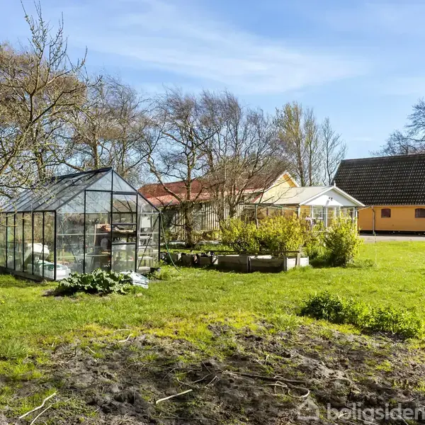 Et drivhus står i en grøn have ved siden af blomsterbede. I baggrunden ses flere bygninger med røde og gule facader, omkranset af træer under en blå himmel.