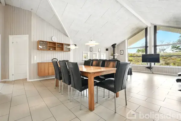 Et spisebord med otte sorte stole står i en lys stue med højt til loftet. Store vinduer bagved giver udsigt til landskabet, mens en reol og fjernsyn også ses.