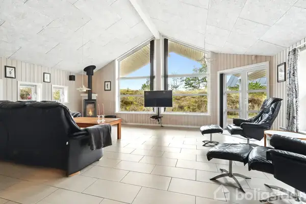 Skindlædermøbler står i en stue med store vinduer, der giver udsigt til en græsmark. En brændeovn er placeret i hjørnet og et fjernsyn hænger på væggen.