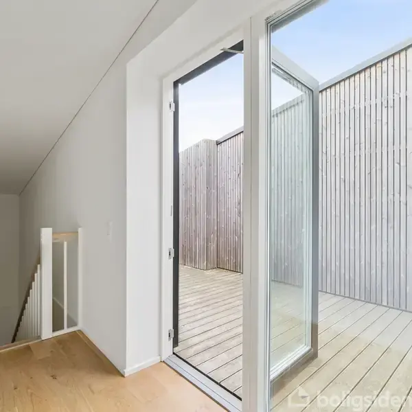 En åbnet glasdør i en lys gang viser en træterrasse omgivet af høje hegn. Trægulve og hvidmalede vægge præger det moderne, minimalistiske interiør. Ingen tekst til stede.