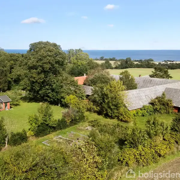 En gård med røde murstensbygninger og grå tage ligger omkranset af frodig natur nær vandoverfladen. En have med et lille skur ses til venstre, skov omringer området.