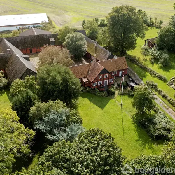 En rød bindingsværksgård omgivet af frodige træer og grønne marker. En grusvej fører til gården, der ligger i et landligt område med yderligere bygninger i baggrunden.