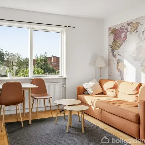 En sofa står i et hjørne ved siden af et verdenskort på væggen. Der er et lille bord foran sofaen og spiseplads ved vinduet, der viser udsigt til grønne træer.