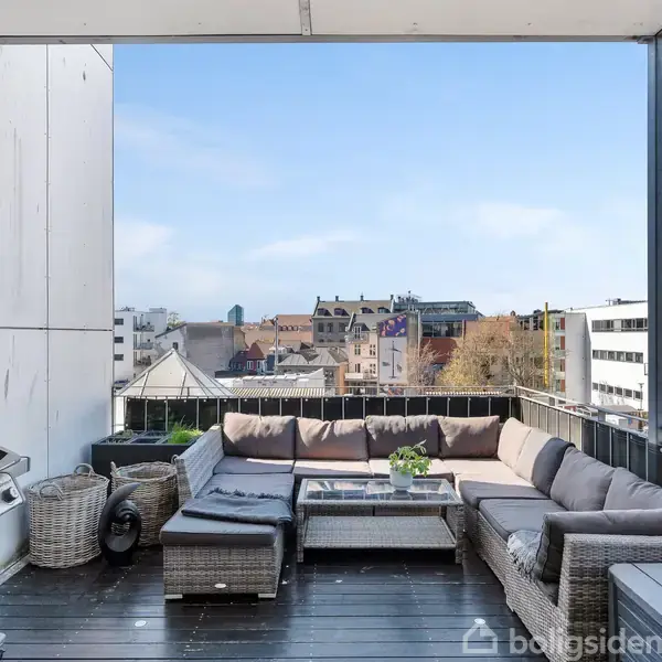 Udendørs terrasse med en grå hjørnesofa omkring et glasbord, omgivet af høje bygninger. En grill står til venstre, og der ses blå himmel over byens tage.