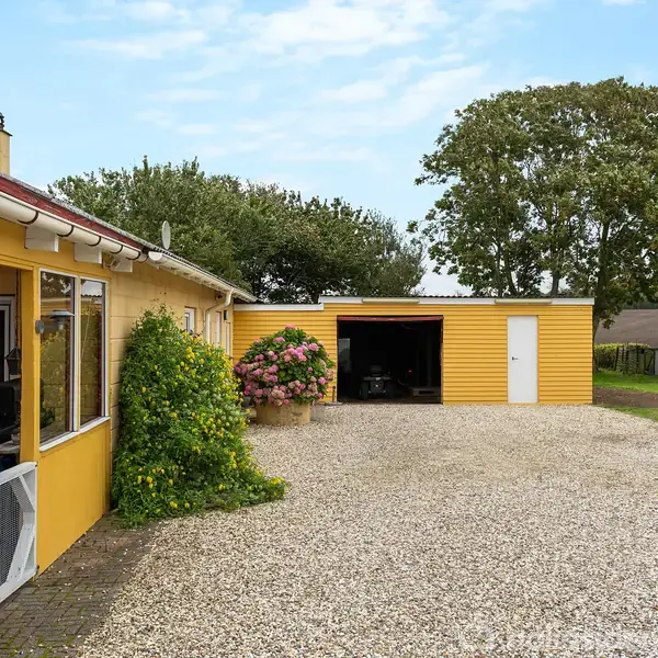 Et gult hus med store vinduer står ved siden af en grusbelagt indkørsel. I baggrunden ses en garage, blomstrende buske og grønne træer.