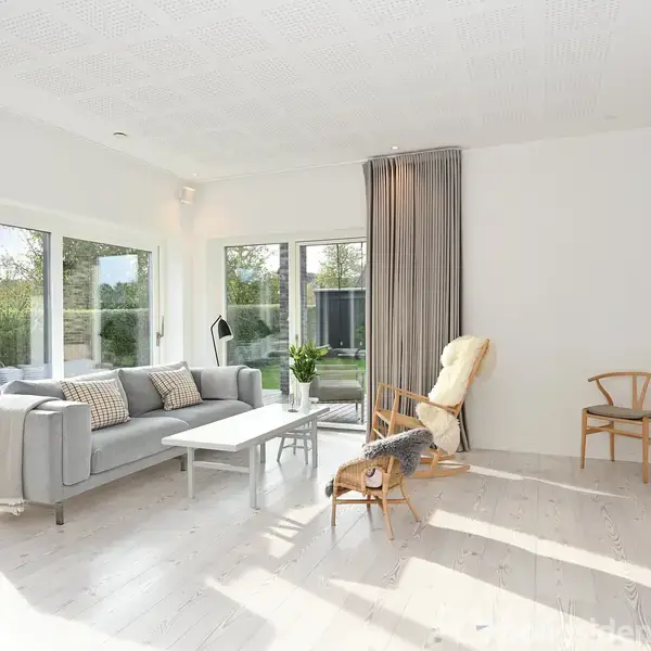 Sofa står i lys stue med store vinduer, der slipper sollys ind. Rummet har lyse møbler, tæppe, lænestol og fladskærm på væggen. Udsigt til have udenfor.