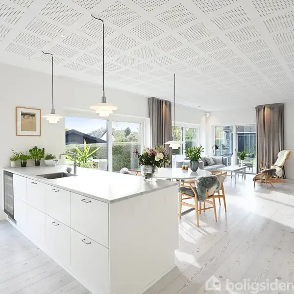 Køkkenø i hvidt står i centrum, omgivet af planter og lamper, i et lyst, moderne rum med store vinduer og udsyn til en have.