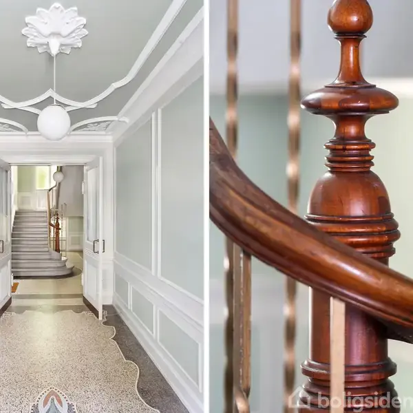 En elegant gang med en udsmykket loftlampe og dekoreret gulv fører til en trappeopgang. Til højre ses en detaljeret, mørk trægelænder langs trappen.