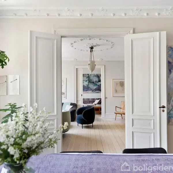 Et elegant, lyst rum med hvide døre åbner ind til en stue med kunstværker og moderne møbler. Et lille træ og blomster på bordet giver liv til omgivelserne.