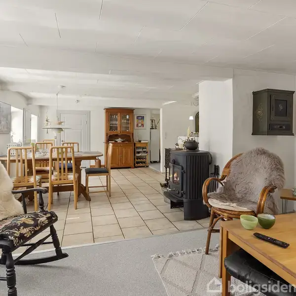 Gyngestol med pels sidder foran sofa i stue med spisebord i baggrunden. En brændeovn står ved væggen. Rummet er lyst med fliser og træmøbler.