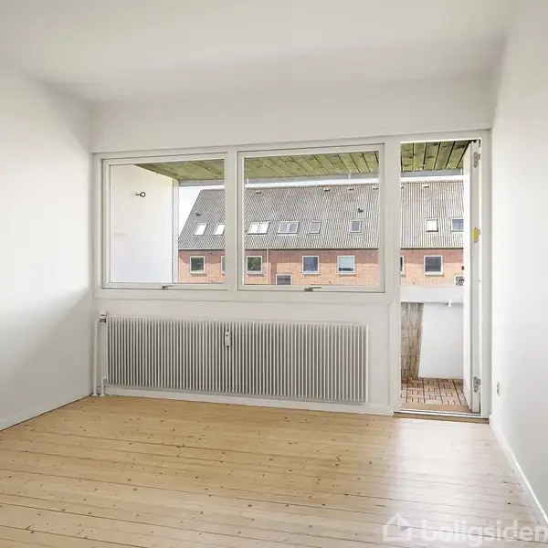 Et tomt værelse med lyse trægulve og hvidmalede vægge har vinduer mod en altan. Udenfor ses en murstensbygning med tagvinduer. Indeklimaet opvarmes af en radiator under vinduet.