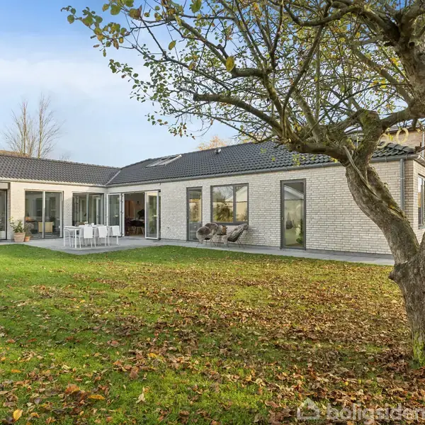 Et moderne enfamiliehus med store vinduer og hvid murstensfacade står i en have med græsplæne og et stort træ. Huset har en terrasse med borde og stole.