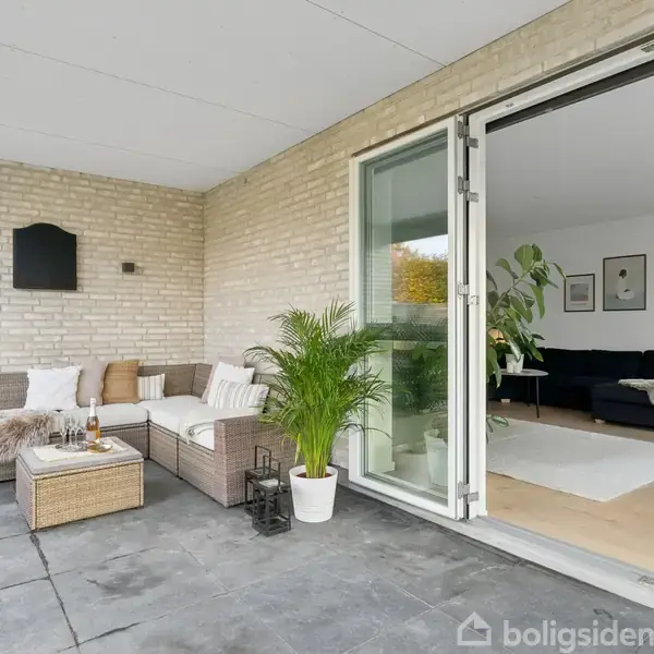 Københavnermøbler er arrangeret på en terrasse, dekoreret med puder og en plante. Skydedøre åbner op til en stue med sofaer og kunst på væggene.