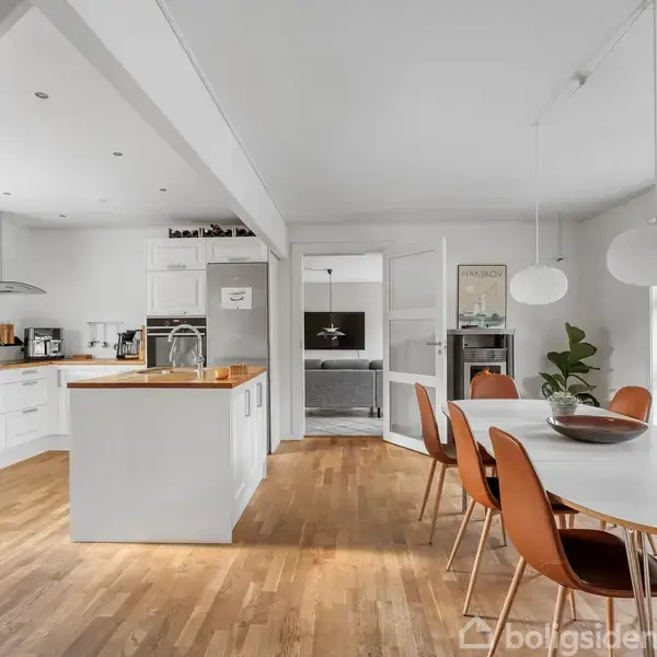 Et moderne køkken-alrum med hvide skabe og træbordplader. Et rundt spisebord med brune stole står til højre. Rummet er lyst og åbent med store vinduer og trægulve.