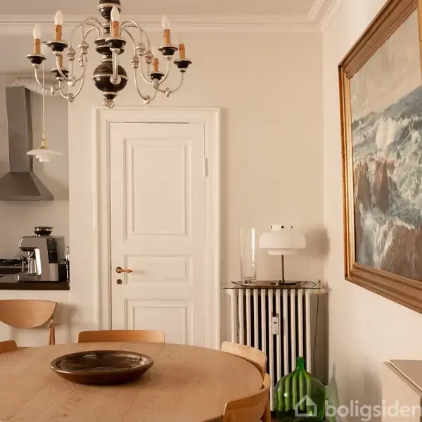 Et rundt træbord står i en spisestue med en lysekrone over sig. Bagved ses en hvid dør, en komfur i et hjørne, og en indrammet havmaleri på væggen.