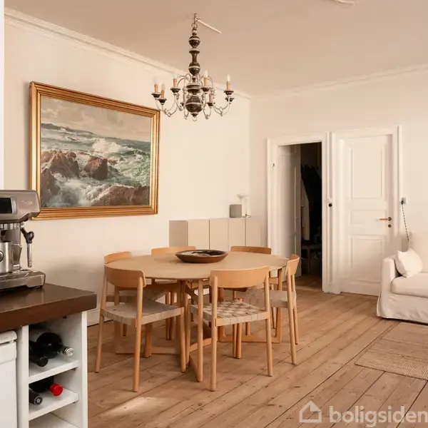 En stue med en rund spisebord i lyst træ omgivet af stole. En espressomaskine står på en køkkenbordplade til venstre. Et maleri hænger på væggen, og en sofa er placeret til højre. En lysekrone hænger over bordet.