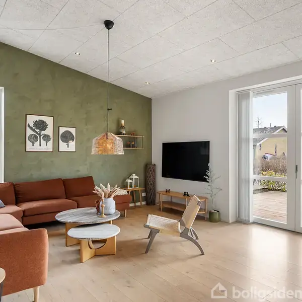 Stue med brun sofa og rundt sofabord, væg-TV og grøn plante. Store vinduer til højre slipper lys ind, moderne indretning med træ- og grønne nuancer. To vægbilleder af træer.