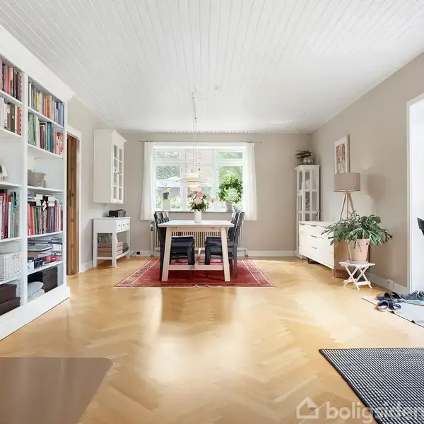 En lys stue med et spisebord i midten, omgivet af stole, står på et rødt tæppe. Reol med bøger og brændeovn til venstre. Store vinduer og dør til højre.